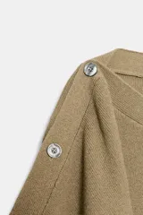 Jersey de punto color beige con cuello barco y mangas anchas tipo capa que presentan detalle de botones a lo largo de los hombros y brazos.