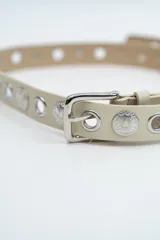 Cinturón de cuero sintético color beige, con hebilla metálica plateada y diseño decorado con ojales metálicos y apliques circulares grabados a lo largo de toda la tira.