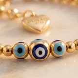 Pulsera elástica de cuentas doradas con baño de oro, intercaladas con dijes de ojos turcos en tonos azul, blanco y negro.