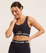 Top deportivo negro de microfibra con escote redondo y abertura tipo cutout en el centro del busto. Posee copas desmontables y una banda elástica ancha bajo el busto con el texto "KEEP MOVING" repetido en blanco.