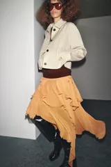 Trench corta color beige con cuello solapa, manga larga con trabilla y botón en los puños, bolsillos de vivo en el delantero y cierre frontal con botones oscuros.