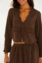 Blusa blanca de manga larga con escote en V, detalles de volados horizontales y cierre frontal con tiras ajustables.