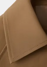 Chaqueta corta de algodón color marrón tostado, con cuello alto, cierre frontal de botones, bolsillos ribeteados y mangas largas con trabillas ajustables en los puños.