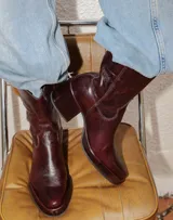 Bota estilo tejano de cuero color bordó con acabado brillante, taco cuadrado y punta cuadrada. Presenta costuras decorativas en el empeine y caña media.