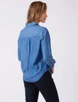 Camisa de tencel azul, marca Weekend Routine, con cuello solapa, botones metálicos frontales y manga larga regulable con presilla y botón. Presenta canesú horizontal en la espalda con tablón central y bajo asimétrico redondeado.