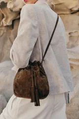 Bolso tipo bandolera de formato mini, confeccionado en piel de ante color marrón. Presenta un diseño de saco con cierre mediante cordón fruncido y detalle de borlas. Incluye una correa larga ajustable y herrajes metálicos dorados.