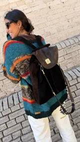 Mochila Brassy confeccionada en combinación de gamuza marrón oscuro y cuero negro, con solapa de cuero texturado y herrajes metálicos en tono oro viejo. Presenta correas regulables y diseño funcional para el día a día.