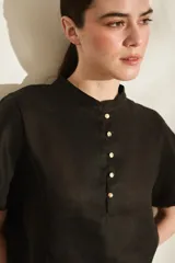 Blusa negra de lino con cuello mao, mangas cortas abullonadas y cierre frontal parcial con botones dorados. Presenta pequeños tajos laterales en el ruedo.