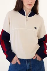 Sweater de punto acanalado con cuello alto y cierre frontal parcial. Presenta un diseño tricolor en bloques con cuerpo color crema, cuello y puños azul marino, y detalles en rojo en las mangas. Incluye una pequeña etiqueta con logo en el pecho.