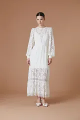 Vestido largo de encaje color blanco, con mangas largas abullonadas, cuello redondo y falda con volados en el ruedo. Presenta un diseño con transparencias y forro interno.