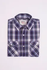 Camisa de manga corta Mistral con estampado de cuadros en tonos violeta, azul, blanco y negro. Presenta cuello clásico, cierre frontal con botones a presión y dos bolsillos delanteros con solapa y botón.
