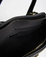 Cartera tipo tote bag color negro de símil cuero con textura granulada, asas de mano y correa bandolera ajustable. Cuenta con herrajes dorados envejecidos y tira decorativa frontal con hebilla.