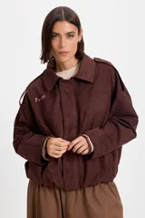 Campera bomber de gamuza color chocolate con cuello de corderito sintético desmontable en color crema. Presenta un diseño holgado con cierre frontal, hombreras decorativas y bordado sutil en el pecho.