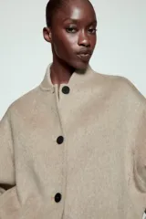 Cazadora bomber beige confeccionada en hilatura con lana, con cuello subido, manga larga y bolsillos frontales. Presenta cierre frontal con botones oscuros y puños, cuello y ruedo en tejido rib.