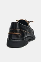 Zapato tipo náutico de piel color negro, con detalle de pespuntes en contraste, cordones decorativos y puntera redonda con costura fruncida.