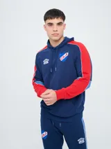 Canguro deportivo Umbro del Club Nacional de Football, modelo Nexo para hombre. Diseño en color azul marino con mangas rojas y detalles en celeste y blanco, capucha con cordón ajustable y bolsillo tipo canguro.