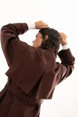 Trench color chocolate con cuello alto, cierre frontal con botones y cinturón ajustable en la cintura. Presenta mangas largas con puños ajustables mediante tiras y hebillas, y bolsillos frontales.