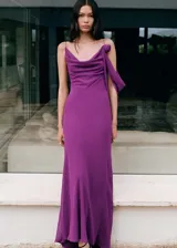 Vestido largo de fiesta color violeta, con diseño recto, cuello drapeado y tirantes finos. Presenta un detalle de flor con drapeado en uno de los hombros y cierre de cremallera lateral.