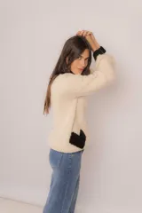 Buzo tejido a mano en lana merino, color crema con detalles en negro en el cuello, puños y un diseño de corazón en la parte inferior. Presenta un corte amplio y mangas largas.
