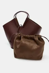 Bolso tipo shopper con forma de capazo, color marrón oscuro, confeccionado en material liso con pespuntes visibles en el cuerpo. Posee doble asa de mano y un asa bandolera ajustable. Incluye un bolso interior extraíble que se cierra mediante fruncido y un cierre principal con botón imantado.