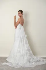 Vestido de novia blanco strapless con corsé armado y falda corte sirena de tul drapeado. Presenta apliques de flores de organza en la falda y una cola romántica de tul.