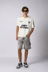 Remera tipo chomba de tejido de punto color blanco, con cuello polo y diseño de rayas verticales sutiles. Presenta estampado gráfico frontal con texto y logo, además de detalles de rayas negras en los hombros.