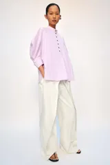Camisa confeccionada en hilatura de algodón de color rosado claro, con cuello mao y cierre frontal parcial con botones a contraste. Presenta mangas por debajo del codo abullonadas y bajo asimétrico con aberturas laterales.