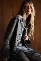 Campera estilo militar napoleón confeccionada en cuero ovino, con cierre frontal y solapa decorativa con botones metálicos y alamares de gamuza en tono crudo. Cuenta con cuello camisero, pespuntes visibles y diseño estructurado de manga larga.
