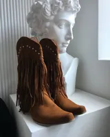 Botas texanas de gamuza color marrón, con flecos que cubren la caña y detalles de tachas plateadas en la parte superior.