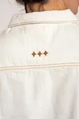 Chaleco de denim blanco con diseño sin mangas, cuello camisero, bolsillos frontales con solapa y cierre de botones metálicos con detalle de ribete ondulado en contraste.