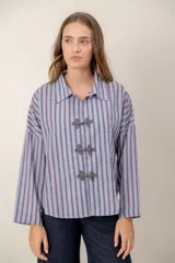 Camisa de corte recto y holgado con estampado de rayas verticales en tonos azul y rojo. Presenta cuello clásico y cierre frontal mediante nudos decorativos tipo alamar en color gris.