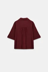 Camisa negra de textura arrugada, con cuello solapa, escote en V y cierre frontal de botones. Cuenta con mangas cortas terminadas en vuelta con trabilla y botón.