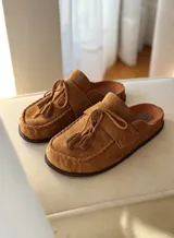 Zueco tipo mocasín destalonado confeccionado en gamuza color tabaco, con detalle de cordones y borlas en el empeine y costura reforzada en el contorno de la puntera.