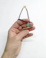 Collar corto compuesto por pequeñas cuentas facetadas de color bordó y un dije colgante circular de piedra verde con detalle dorado en el centro.