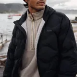 Campera puffer negra con capucha, confeccionada en poliéster con relleno reciclado. Presenta un diseño acolchado con costuras curvas, cierre frontal con cremallera y bolsillos delanteros.