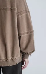 Buzo de cuello redondo color marrón claro, confeccionado en algodón premium con textura acanalada. Presenta costuras expuestas y detalles deshilachados en cuello, puños y ruedo, con un calce oversize y mangas abullonadas.