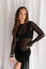 Vestido corto negro de encaje transparente, con mangas largas y falda ajustada con detalle lateral asimétrico tipo pareo.