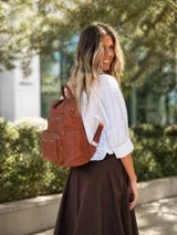 Mochila de símil cuero color suela, con apertura superior, tres bolsillos frontales con cierres metálicos y bolsillos laterales. Incluye correas ajustables para la espalda y una correa larga de galón para usar como bandolera.