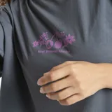 Remera de corte oversize color gris, con cuello redondo y mangas cortas. Presenta un estampado pequeño en el centro del pecho con motivos florales y de frutas en color rosado.
