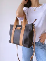 Cartera de cuero negro con detalles en cuero color suela, incluyendo asas fijas y correa larga ajustable. Presenta una etiqueta frontal con logo grabado y cierre superior.