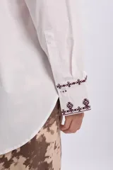 Camisa blanca de corte oversize, con mangas largas y puños anchos decorados con bordados en tono oscuro. Cuenta con cuello camisero clásico y cierre frontal de botones.