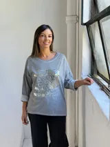 Remera gris de manga larga con cuello redondo y diseño holgado. Presenta un aplique frontal rectangular con detalles de lentejuelas plateadas y estampado tipográfico.