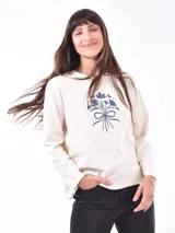 Remera de manga larga color crema con cuello redondo y un estampado central de un ramo de flores rosadas y naranjas en un jarrón azul oscuro.