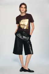 Camiseta de algodón color marrón oscuro, de corte regular, cuello redondo y manga corta. Presenta una estampación metalizada dorada en el parte frontal con texto "REYKJAVIK ART GALLERY" y "ABSTRACT MOVEMENTS Reykjavik, Iceland, 1975".