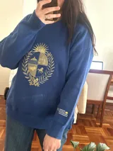 Buzo de tejido de punto color azul, con cuello redondo y diseño holgado. Cuenta con un gran bordado dorado en el pecho que representa el escudo patrio rodeado de laureles.