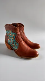 Botas de cuero color marrón tierra con taco medio, detalles de costuras artesanales en el borde superior y un aplique lateral con diseño geométrico pintado en tonos turquesa y cobrizo.