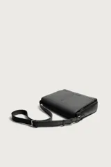 Bandolera de diseño rectangular en color negro con acabado texturizado. Cuenta con una solapa frontal con detalle metálico, cierre imantado y asa de hombro ajustable. El interior incluye un bolsillo plano y otro con cierre, además de un bolsillo plano adicional en la parte frontal.