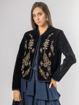 Chaqueta de terciopelo negro con bordados florales y ornamentales en tonos dorados, rosados y azules. Presenta un corte recto, cuello redondo y cierre frontal abierto con detalles de costuras decorativas en los bordes.