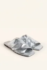 Sandalia tipo slide de cuero vacuno color blanco, con diseño de tiras cruzadas y fruncidas. Presenta una puntera cuadrada y una suela plana de cuero con un pequeño taco de 2 cm.