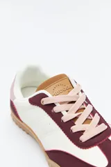 Zapatilla urbana de mujer con silueta retro, combinando paneles de cuero blanco y gamuza gris claro. Posee un talón trasero en gamuza negra y suela robusta de goma color caramelo con relieve.